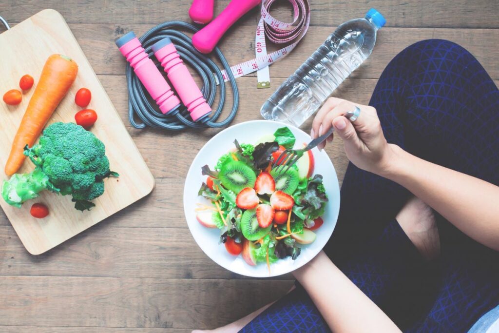 A woman is sitting just out of frame. She is holding a bowl of salad with fruits in it. Veggies and workout equipment are beside her.