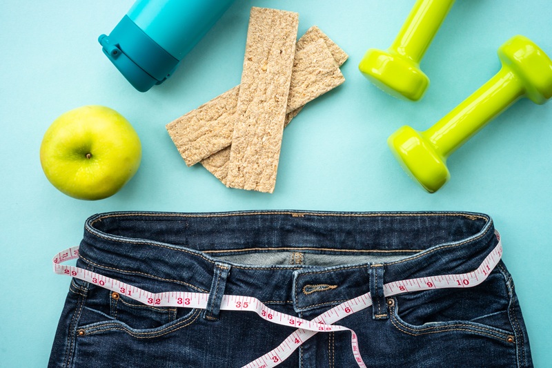 A light blue/teal background. Jeans with measuring tape through the belt loops. Bottle of water, lime green dumbbells, a green apple, and crackers are scattered around.