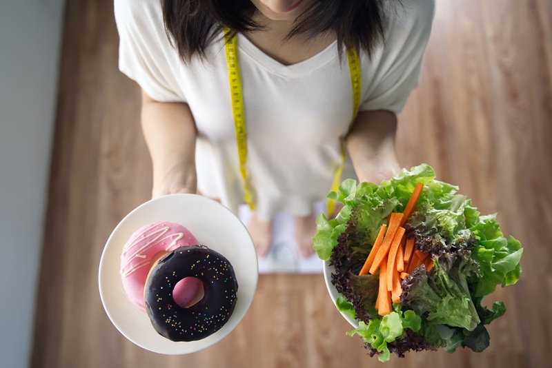 Top view of young Asian girl holding dishes of vegetable salad and donuts standing on weight measurement scale with measure tape on neck. Decision making for her health.